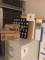 Assorted office supplies on filing cabinet including box with writing instruments, binders, letter board and photo paper