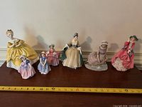 Group shot of seven Royal Doulton figurines on table with ruler for scale
