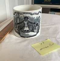 Front of white ceramic cup showing black transferware illustration and scale reference