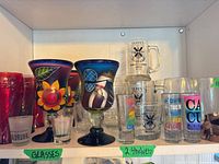 Overview of assorted glassware on two shelves, shows Mexican goblets, Queen’s beer mugs, printed tumblers, shot glasses
