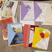 Top-down view of assorted greeting cards showing designs such as bridal couple, musical Father’s Day, floral iris, purple lily wife card, red foil scroll, balloon TLC etc.