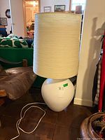 Full view of lamp showing white ceramic base, beige fabric drum shade, power cord