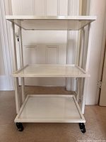 Front view of white metal trolley cart showing three glass shelves and caster wheels