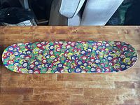 Full view of skateboard deck showing colourful cartoon face pattern and absence of hardware