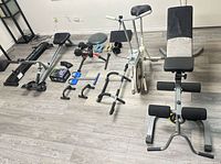Wide view showing exercise bike, bench, rowing machine, dumbbells, push-up bars, ab roller, balance disc and accessories arranged on floor