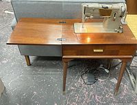 Singer sewing machine set into wooden cabinet with extension leaf opened, foot pedal below
