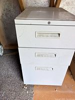 Front view of white 3-drawer metal filing cabinet showing handles, lock and surface wear