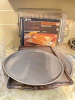 Stack of three steel cookie sheets, round pizza pan, microwave bacon rack with packaging visible