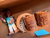 Shelf view showing two carved Bahamas mugs, wood moon with beads, folk art doll