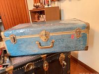 Front view of blue/teal suitcase trunk on top of another trunk showing textured exterior and brass hardware