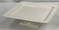 Cream square ceramic pedestal cake stand showing beaded rim and base