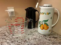 Pyrex glass measuring cup, black plastic carafe, white carafe with pear design on countertop