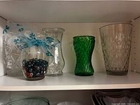 Shelf view showing five vases including clear pressed glass, flared vase, jar with marbles, green twisted vase, smoke diamond-point vase