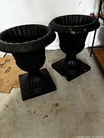 Two black plastic pedestal urn planters shown side by side on concrete floor