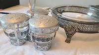 Three small condiment jars with lids and spoons beside metal casserole stand