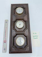 Full view of wooden plaque with three brass-rimmed dials next to ruler for scale
