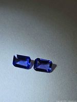 Pair of rectangular emerald-cut violet-blue gemstones on neutral background
