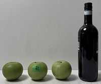 Three green-patinated bronze apples beside a wine bottle for size reference