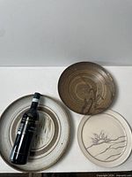 Group view of three signed shallow pottery plates with wine bottle for scale