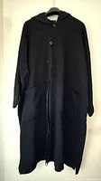 Front view of full-length black coat on hanger showing hood, buttons, patch pockets