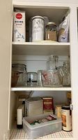 Cabinet shelf with tins, canisters, jars, pitcher, measuring cup, coffee grinder, tea box and plastic bin