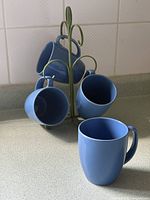Group of four blue mugs displayed on green metal tree with one mug in foreground