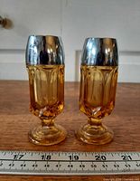 Full view of both amber shakers side by side showing pedestal bases and chrome-look lids with ruler for scale