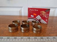 Six brass napkin rings lined in front of original box with ruler for scale