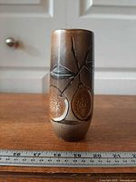 Full front view of slender brown ceramic vase with fruit motif beside ruler for scale