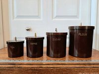Front view of four graduated dark brown plastic canisters labeled TEA, COFFEE, SUGAR, FLOUR with gold-tone knobs