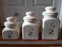 Front view of three graduated white ceramic canisters with rose motif