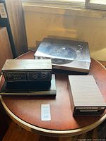 Four electronic items on round table: turntable, clock radio, Blu-ray player, 8-track unit