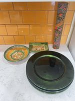 Stack of four green tinted glass chargers beside two glazed ceramic plates and decorative tube