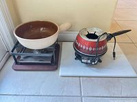 Two fondue sets on white slab: beige ceramic pot on metal stand with burner and Oster red electric fondue pot