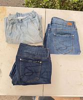 Three folded pairs of jeans on table: two Seven7 medium-dark, one light wash GU