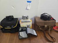Overview of three cameras, Nikon bag, Kodak box, brown case on table