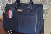 Front view showing bag, handles, shoulder strap, exterior pocket, brand plaque