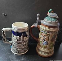 Front view of both steins showing Heidelberg and hunting designs, pewter lids visible