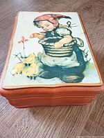 Top view showing cream metal panel with girl feeding chicks illustration on wooden music box