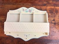 Front view of cream wall shelf showing cubbies, perforated panel, hooks, floral motif