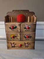 front view showing five labeled drawers, painted flowers, red knobs, spool dowels and tomato pin cushion
