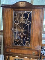 Front view showing wood cabinet, glass door with metal scrollwork, lower drawer, turned legs