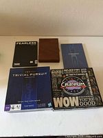 Five items: Trivial Pursuit box, Cranium WOW box, closed wooden backgammon case, black Fearless album, blue album