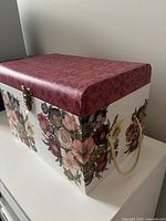 Closed floral storage box, showing lid pattern, latch and one rope handle
