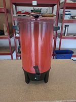 Front view of orange and black electric coffee urn showing spigot, handles, and base