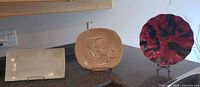 Three plates on countertop with stands: rectangular ceramic platter, tan pottery car-relief plate, round red/black resin plate