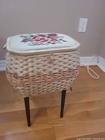 Closed wicker sewing basket on black legs, embroidered lid visible