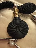 Close-up of perfume atomizer pendant showing matte black bottle, gold collar, squeeze bulb, part of twisted cord