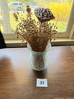 Clear pressed glass vase with dried seed pods and stems on wood surface