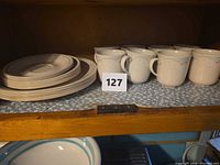 Stack of dinner and dessert plates beside eight mugs on shelf